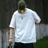 Person in Soft Boy Aesthetic outfit: oversized white tee, green cap, necklace, and black cargo pants, holding a bag against a blurred outdoor background. DAXUEN Aesthetics streetwear style.