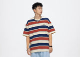 A young man wearing a red, blue, and beige striped T-shirt, light blue jeans, and a silver chain necklace stands against a plain white background. He has curly hair and accessorizes with a wristwatch and bracelet, showcasing a casual streetwear style