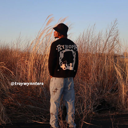 Black RYHOPM graphic sweater, light blue cross-designed jeans, black cap, outdoor field background
