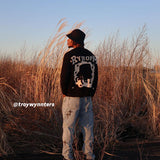 Black RYHOPM graphic sweater, light blue cross-designed jeans, black cap, outdoor field background